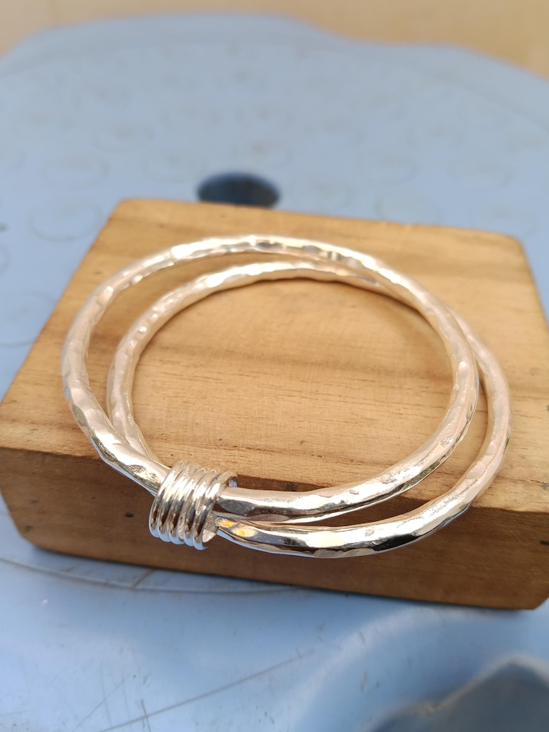 May include: A silver bangle bracelet featuring two interlocking circular bands with a hammered, textured finish. The two bands are joined together by a small, smooth, cylindrical silver connector piece that holds them in a fixed, overlapping position.