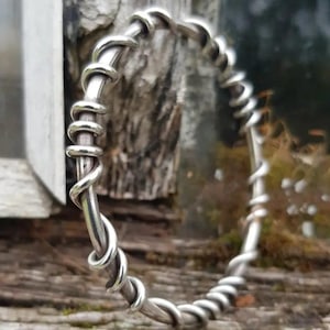 May include: A silver-toned bangle bracelet with a twisted wire design. The bracelet is made of a thick, round metal and features a continuous spiral pattern. The design gives the bracelet a rustic, handcrafted appearance. The bracelet is shown outdoors.
