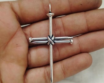 May include: A silver cross pendant made from nails. The nails form the cross shape, with a knot securing the intersection. The pendant has a small loop at the top for a chain. The cross is held in a hand.