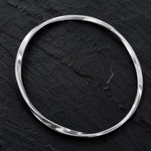 Puede incluir: Un brazalete de plata pulida con un diseño de metal retorcido continuo. La banda circular tiene una superficie lisa y reflectante que capta la luz, creando reflejos sutiles a lo largo de los bordes curvos de la estructura metálica.
