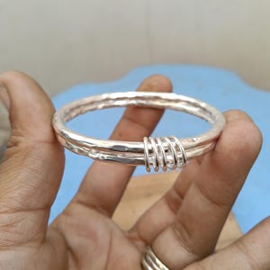May include: A silver-colored bangle bracelet featuring a hammered, textured surface. The bracelet is composed of two circular bands joined together by four smaller, interconnected silver-colored rings that wrap around both main bands.