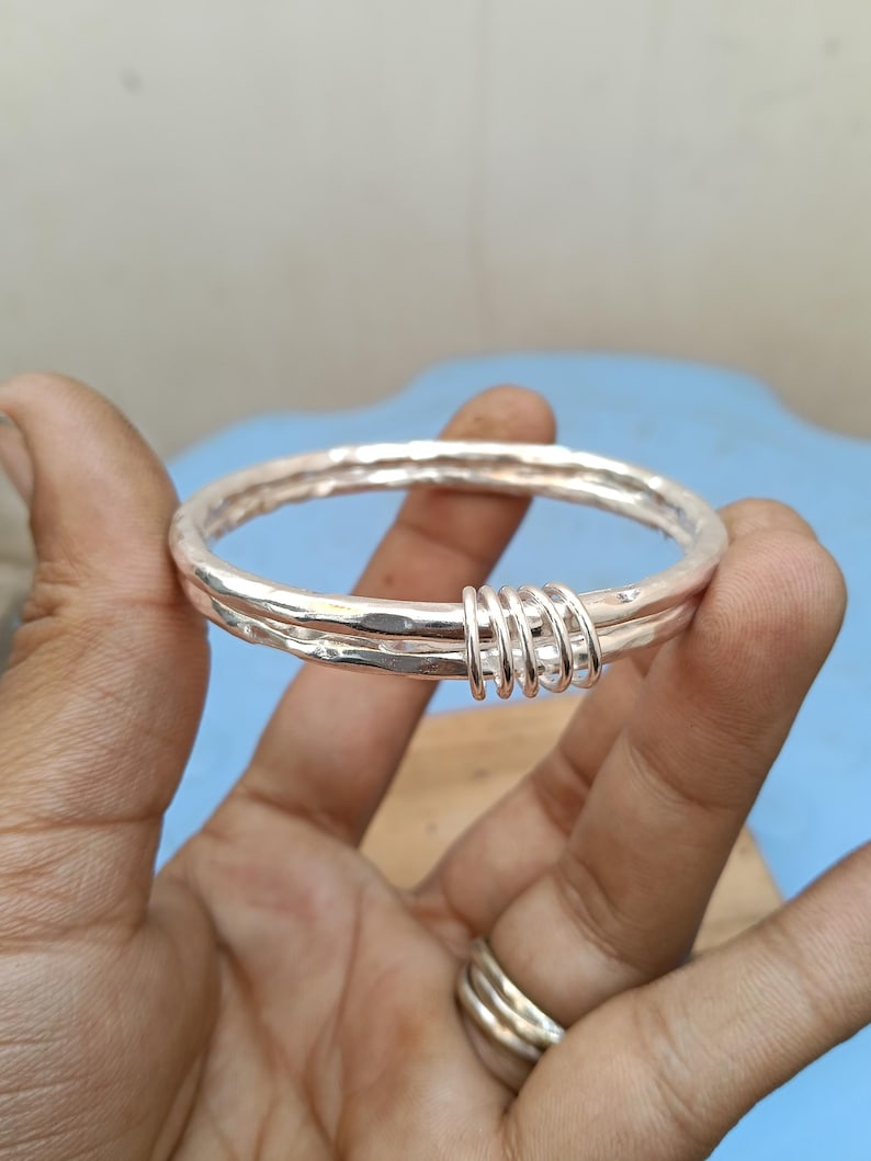 May include: A silver-colored bangle bracelet featuring a hammered, textured surface. The bracelet is composed of two circular bands joined together by four smaller, interconnected silver-colored rings that wrap around both main bands.