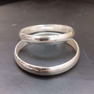 May include: Two silver bangles with a rounded profile are stacked on a dark surface. The bangles have a smooth, polished finish, reflecting light. The simple design suggests a classic jewelry style.