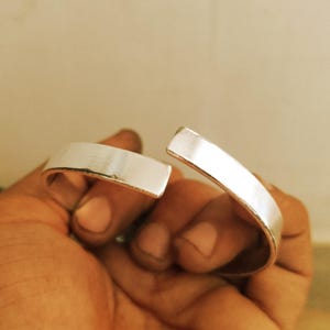 May include: A silver-colored cuff bracelet with a rectangular design. The bracelet is open, revealing the inner curve. The metal has a polished, reflective surface. The bracelet is held in a hand.