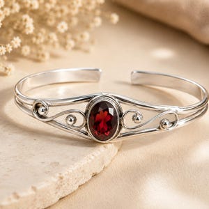 May include: A silver cuff bracelet featuring a central oval-cut deep red garnet gemstone. The metal band displays intricate wire-work scroll patterns on both sides of the stone, with small silver spheres accenting the ends of the decorative swirls.