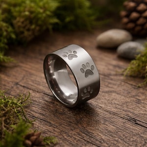 May include: A silver-colored ring with paw print designs. The ring is wide and has a polished finish. The paw prints are evenly spaced around the band. The ring is set against a wooden surface with moss and stones.