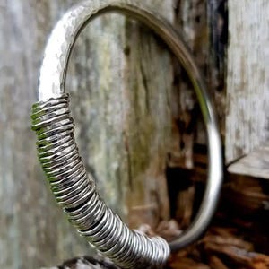Puede incluir: Un brazalete de plata con una textura martillada en la parte superior y un diseño de bobina enrollada en la parte inferior. El brazalete se muestra sobre un fondo de madera envejecida.