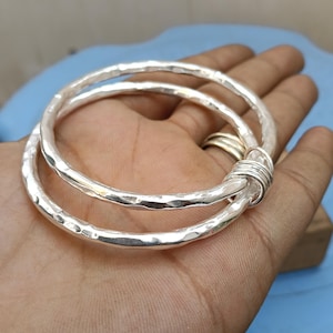 May include: Two silver-colored bangles with a hammered, textured finish. The two circular bracelets are linked together by a small, tightly coiled silver-colored wire wrap that secures them in a fixed, overlapping position.