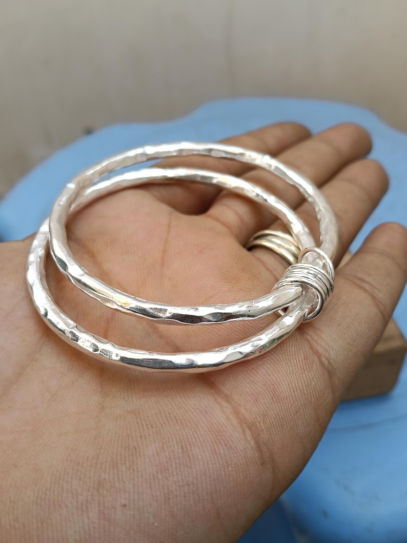 May include: Two silver-colored bangles with a hammered, textured finish. The two circular bracelets are linked together by a small, tightly coiled silver-colored wire wrap that secures them in a fixed, overlapping position.
