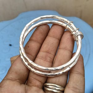 May include: A silver bangle bracelet featuring a hammered, textured surface. The design consists of two overlapping circular bands that are joined together at one side by a series of small, coiled silver wire loops, creating a layered appearance.