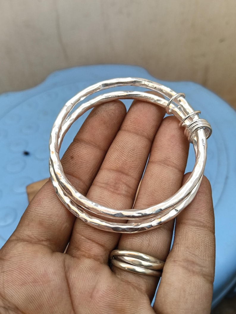 May include: A silver bangle bracelet featuring a hammered, textured surface. The design consists of two overlapping circular bands that are joined together at one side by a series of small, coiled silver wire loops, creating a layered appearance.