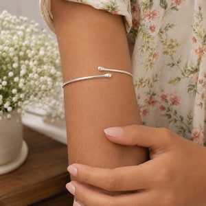 Può includere: Un bracciale aperto color argento caratterizzato da un sottile cinturino liscio che termina con due piccole estremità arrotondate a forma di sfera. Il bracciale è indossato sull'avambraccio e presenta un design semplice e lineare.