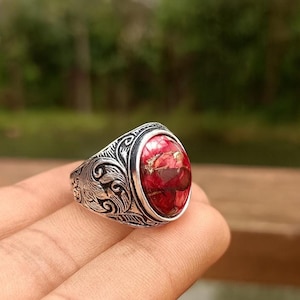 Può includere: Un anello d'argento con una gemma rossa ovale. La gemma ha un motivo unico di rosso e oro. La fascia dell'anello presenta un design intricato e inciso. L'anello è mostrato sul dito di una persona.