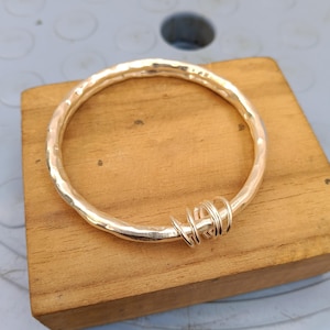 May include: A circular gold-toned bangle bracelet with a hammered, textured surface. The bangle features a decorative section consisting of several small, thin gold-toned wire loops wrapped tightly around one side of the main circular band.