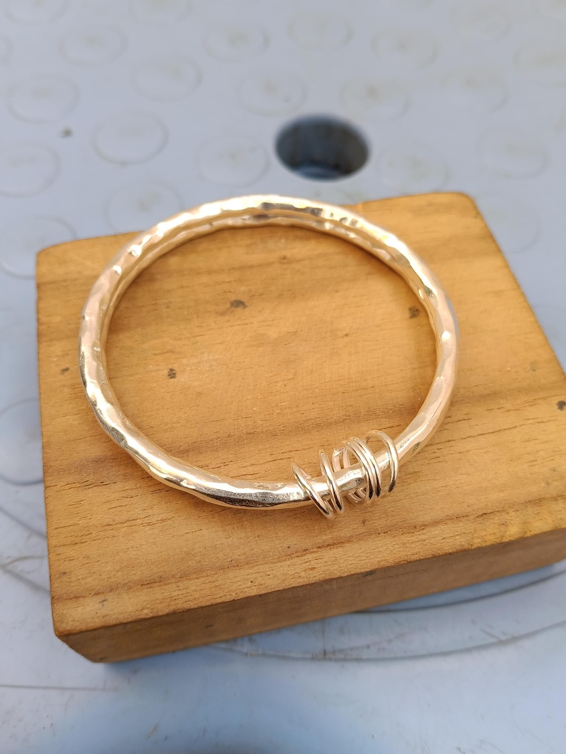 May include: A circular gold-toned bangle bracelet with a hammered, textured surface. The bangle features a decorative section consisting of several small, thin gold-toned wire loops wrapped tightly around one side of the main circular band.