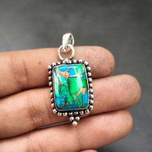 May include: A rectangular pendant with a vibrant turquoise, green, and copper-colored stone set in a silver-toned frame. The frame has a beaded border and a loop for a chain. The pendant is held in a hand.