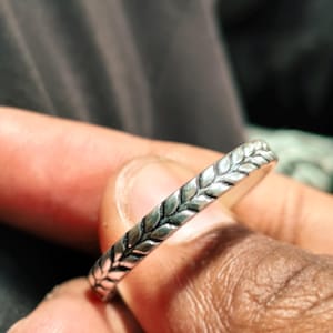 Könnte beinhalten: Ein silberner Ring mit einem Blattmuster. Der Ring ist schmal und hat ein sich wiederholendes Design aus Blättern entlang seines Bandes. Der Ring wird zwischen den Fingern gehalten, wobei der Fokus auf den Details und der Textur liegt.
