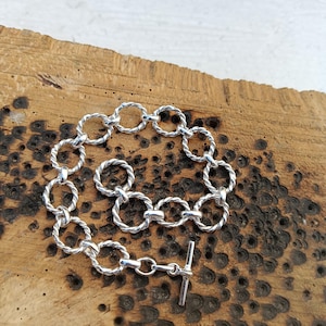 Puede incluir: Un brazalete plateado con una serie de círculos entrelazados, trenzados como cuerdas. El brazalete tiene un cierre de palanca para asegurar el ajuste. La joya se exhibe sobre una superficie de madera texturizada.