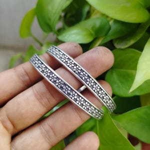 Puede incluir: Dos brazaletes plateados con un patrón floral se sostienen en una mano. Los brazaletes tienen un diseño detallado de pequeñas flores muy juntas. El fondo está borroso, con follaje verde.