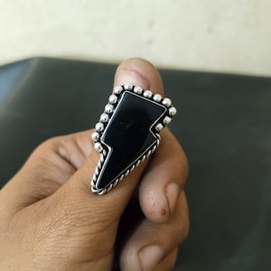 Pode incluir: Um anel de prata com um design de raio preto. O anel apresenta uma pedra preta retangular em forma de raio, rodeada por uma borda de contas. A banda tem um design de corda torcida.