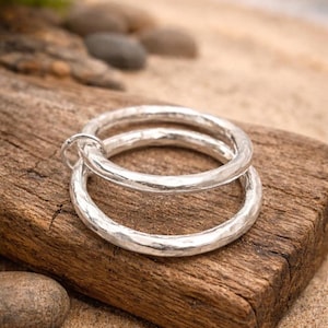 May include: Two silver hammered metal rings, one slightly larger than the other, connected by a small loop. The rings rest on a weathered wooden surface, with a sandy background and small stones.