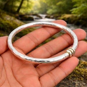Puede incluir: Un brazalete de plata con textura martillada. El brazalete tiene una pequeña sección de anillos de metal dorados. El brazalete se sostiene en una mano, con un fondo natural de río.
