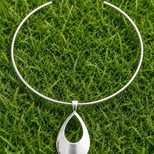 Puede incluir: Un collar plateado con un colgante en forma de lágrima. El collar tiene un diseño de cuello rígido y abierto. El colgante es una forma de lágrima lisa y pulida con un centro hueco. La joya está sobre un fondo de hierba verde.