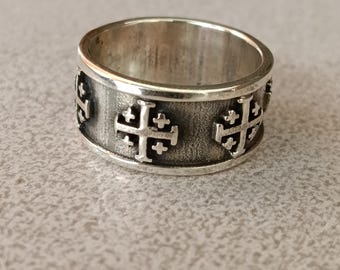 May include: A silver ring with a band design featuring repeating Jerusalem crosses. The ring has a textured surface and a wide band. The crosses are raised and detailed, creating a striking contrast against the ring's surface.