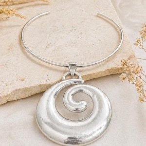 May include: Silver-toned necklace featuring a rigid collar and a large, spiral pendant. The pendant has a textured, hammered appearance. The jewelry is displayed on a neutral-toned surface, with dried plants in the background.
