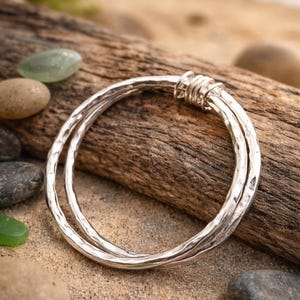 Puede incluir: Una pulsera de plata con textura martillada, compuesta por dos bandas entrelazadas. La pulsera descansa sobre un trozo de madera flotante, rodeada de arena, guijarros y vidrio marino. El diseño de la pulsera es sencillo y elegante.