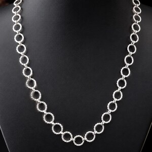 Puede incluir: Un collar de tono plateado que presenta una serie de eslabones circulares interconectados. Cada eslabón individual está elaborado con una textura trenzada similar a una cuerda, creando un patrón uniforme y repetitivo en toda la cadena.