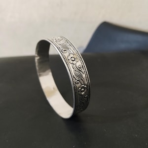May include: A silver-colored bangle bracelet with a floral design. The bracelet features a repeating pattern of flowers, leaves, and beaded accents. The metal has a polished finish and a slightly aged appearance.