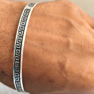 May include: A silver-colored bracelet with a Greek key pattern. The bracelet is a band design and is worn on a wrist. The bracelet is a simple, elegant accessory.