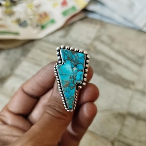 May include: A silver ring with a turquoise lightning bolt design. The turquoise stone is a vibrant blue with brown markings. The ring has a beaded edge and a twisted rope detail around the stone.