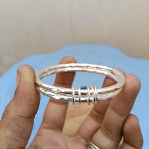 May include: A silver-colored bangle bracelet featuring a hammered, textured surface. The bracelet consists of two connected bands joined together by three small, smooth silver-colored rings that wrap around both main bands, creating a layered look.