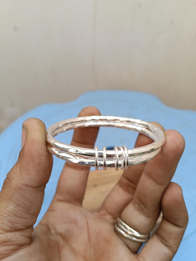 May include: A silver-colored bangle bracelet featuring a hammered, textured surface. The bracelet consists of two connected bands joined together by three small, smooth silver-colored rings that wrap around both main bands, creating a layered look.