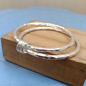 May include: A silver bangle bracelet featuring two interlocking circular bands with a hammered, textured finish. The two bands are joined together by a small, smooth silver coil component that wraps around both sections of the metal wire.