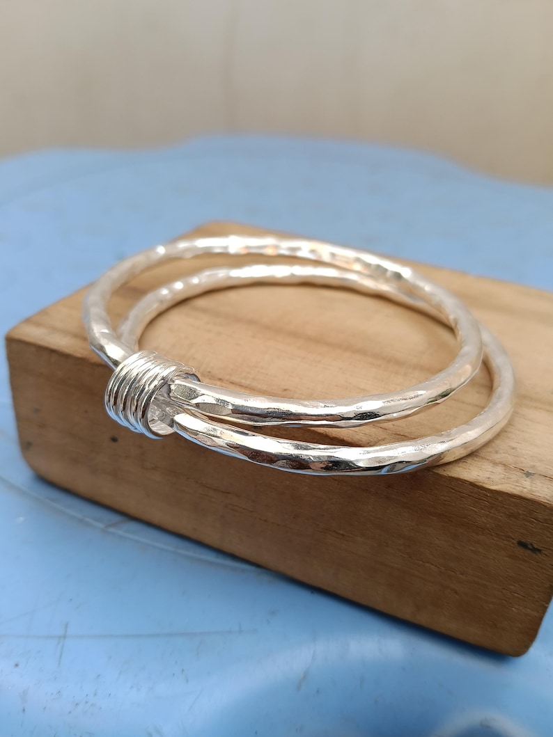 May include: A silver bangle bracelet featuring two interlocking circular bands with a hammered, textured finish. The two bands are joined together by a small, smooth silver coil component that wraps around both sections of the metal wire.