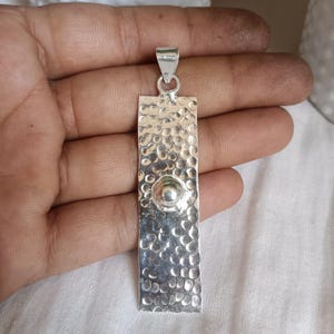 Puede incluir: Un colgante de plata con textura martillada y una pequeña esfera en relieve en el centro. El colgante rectangular tiene un lazo en la parte superior para una cadena. El colgante es de metal brillante y reflectante.