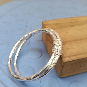 May include: A silver-colored bangle bracelet featuring a hammered, textured surface. The design consists of two overlapping circular bands that are securely joined together at one side by a series of tightly wound silver-colored wire coils.