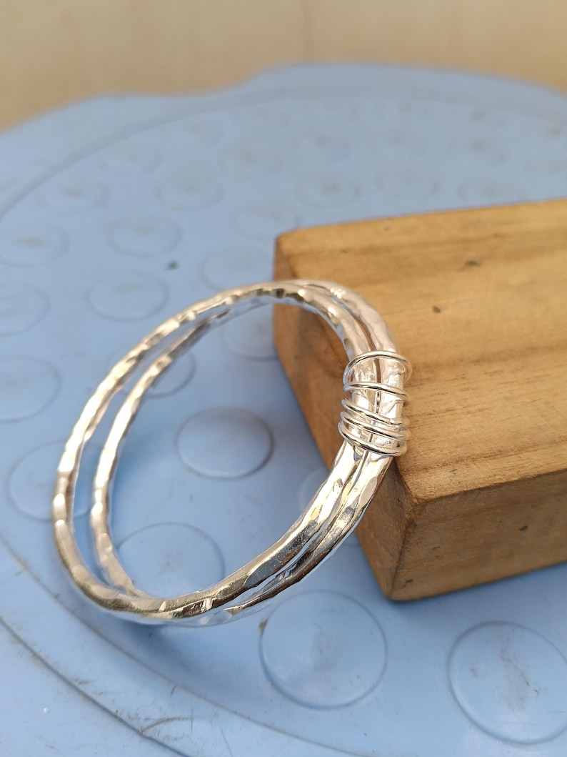 May include: A silver-colored bangle bracelet featuring a hammered, textured surface. The design consists of two overlapping circular bands that are securely joined together at one side by a series of tightly wound silver-colored wire coils.