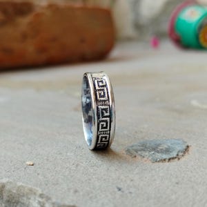 May include: A silver ring with a repeating geometric pattern around the band. The ring is made of a shiny metal and has a detailed design. The ring is displayed on a textured surface.