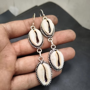 May include: Silver-toned dangle earrings featuring three oval-shaped cowrie shells. Each shell is set in a decorative silver frame with a beaded edge. The earrings have a hook closure.
