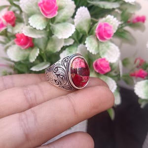 Puede incluir: Un anillo de plata con una piedra preciosa ovalada roja. El anillo tiene un diseño grabado y ornamentado alrededor de la banda. La piedra preciosa tiene una mezcla de colores rojos y dorados. El anillo se muestra frente a un fondo de flores rosas.