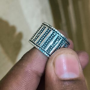 May include: Silver ring with a geometric design. The ring features a series of vertical lines and dotted patterns. The ring is held up by a finger.