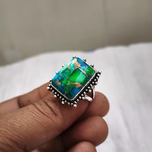 May include: A silver ring featuring a large, rectangular gemstone with a vibrant mix of colors. The stone showcases shades of blue, green, and gold, set in a detailed, ornate silver band. The ring's design is eye-catching and unique.