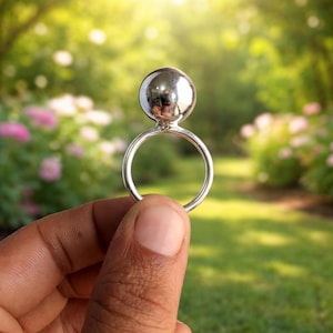 May include: A silver ring with a large, reflective sphere on top. The ring is held up against a blurred background of green foliage and pink flowers, with sunlight shining through the trees. The ring's design is simple and modern.