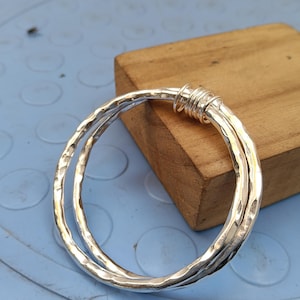May include: A silver bangle bracelet featuring a hammered, textured surface. The design consists of two interconnected circular bands that are joined together at one point by a series of tightly wrapped silver wire coils, creating a layered look.