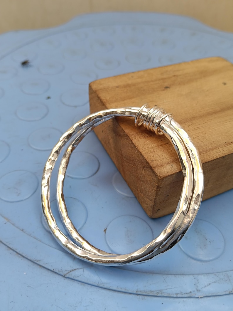 May include: A silver bangle bracelet featuring a hammered, textured surface. The design consists of two interconnected circular bands that are joined together at one point by a series of tightly wrapped silver wire coils, creating a layered look.