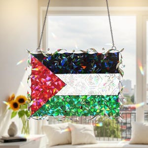 May include: A decorative stained glass panel depicting the Palestinian flag, with red, white, black, and green geometric patterns. The flag is framed with a silver filigree border and hangs from a chain. The panel is displayed in front of a window.